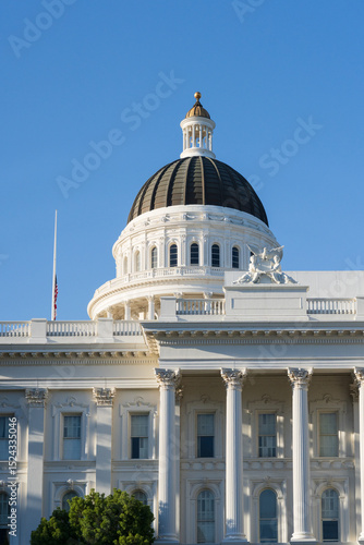 Detailed golden hour vertical view of the California State Capitol building, emphasizing its prominent dome and white columns under a clear blue sky, with subtle greenery and a flag.