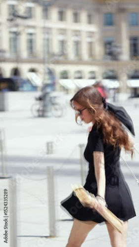 Young woman in a black dress holding a baguette and walking briskly across Place Vendome in Paris on a sunny day, with motion blur capturing energy and fast-paced travel mood