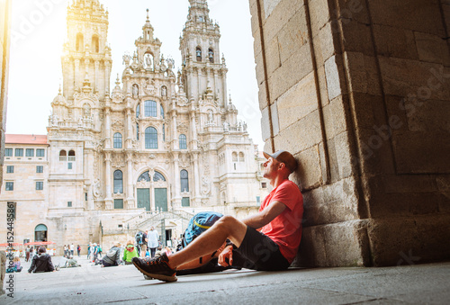 Joyful middle-aged backpacker pilgrim sitting on Obradoiro square in Santiago de Compostela, Spain, with Santiago de Compostela Cathedral — famous Camino de Santiago finish point.