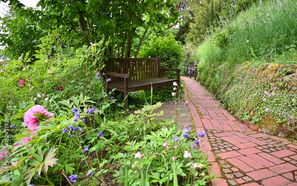 a wooden bench in the beautiful lush green spring garden with pink piones and other spring flowers, Kaufbeuren, Bavaria, Germany a wooden bench in the beautiful lush green spring garden with pink piones and other spring flowers, Kaufbeuren, Bavaria, Germany