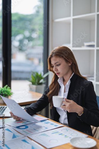 Professional woman reviews financial reports with coffee at her office desk.