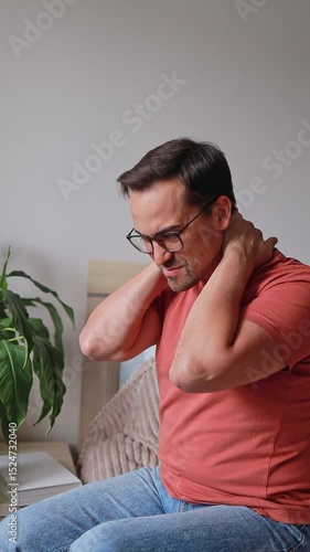 Vertical video of a man suffering from pain and massaging his neck and shoulders while sitting on a bed in the bedroom, osteochondrosis, hernias and protrusions.