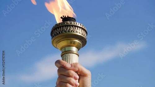 A hand holds a golden torch with a bright flame against a partly cloudy blue sky