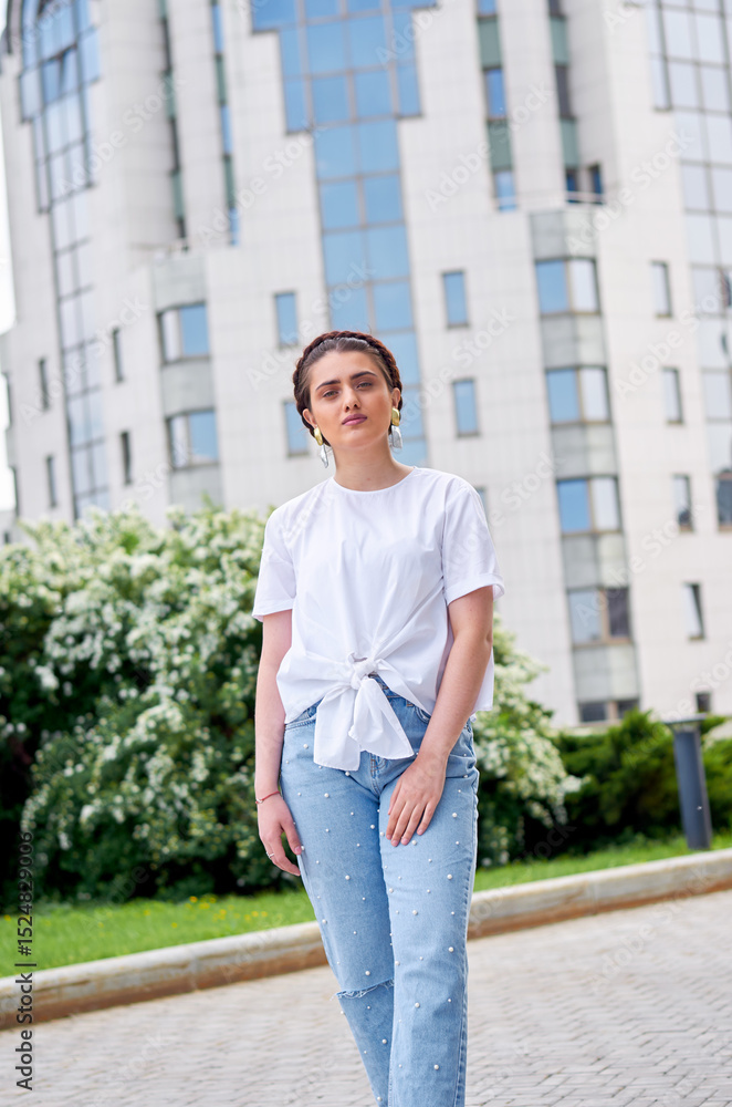 Fototapeta premium A woman in a white shirt and blue jeans walks along the city streets. She has a beautiful athletic figure. The concept of confidence and style. The concept of fashion.