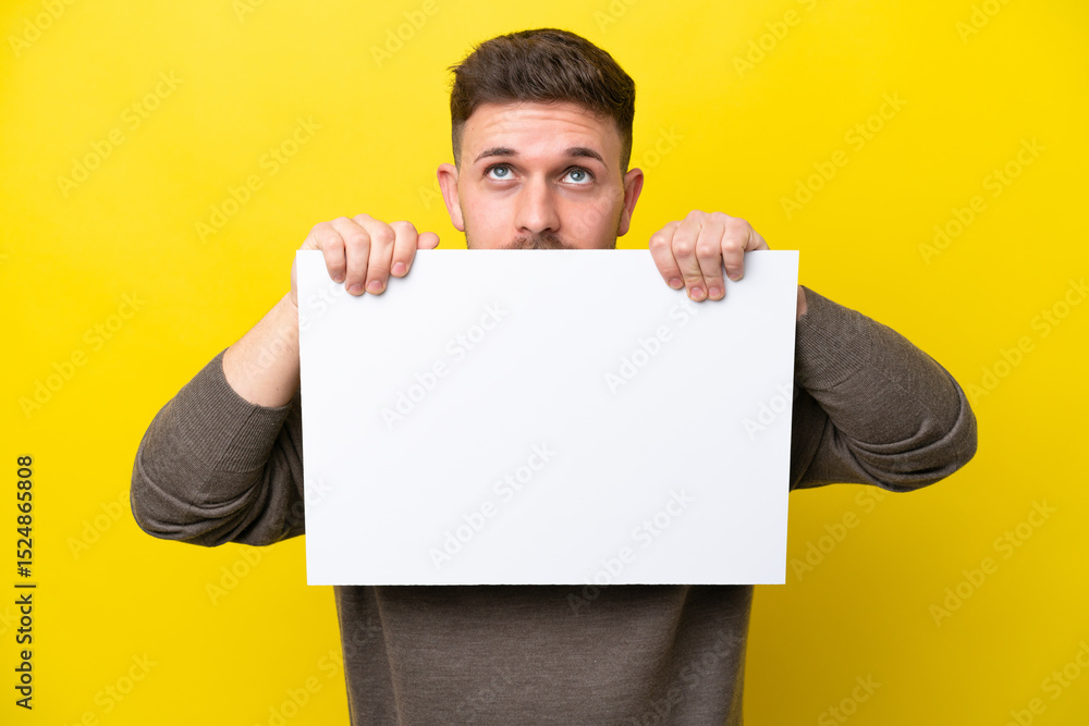 Young caucasian man isolated on yellow background holding an empty placard and hiding behind it Young caucasian man isolated on yellow background holding an empty placard and hiding behind it