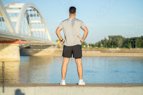 Obraz na plátně Back view to man stands by the water, exuding confidence and tranquility, with hands on hips and a scenic bridge in the background