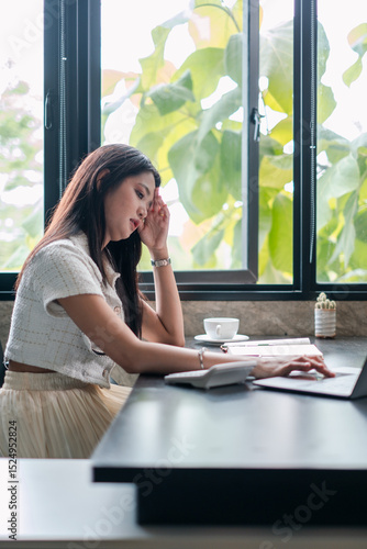 A young woman focuses on her laptop in a stylish cafe, surrounded by natural light and greenery. Free Download A young woman focuses on her laptop in a stylish cafe, surrounded by natural light and greenery. - GFXLoad