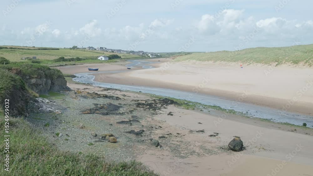A river flow down the picturesque Aberfraw Estuary, Wales, UK, at low tide with sand dunes past boats to a village in the distance under a cloudy sky. A river flow down the picturesque Aberfraw Estuary, Wales, UK, at low tide with sand dunes past boats to a village in the distance under a cloudy sky.