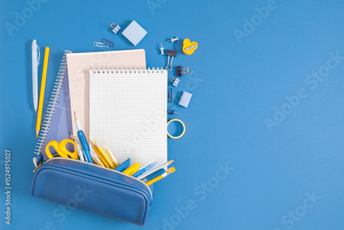 Back to school concept on blue background. Various school stationery items with a pencil case and notebook, as well as a sharpener, paper clips, tape, top view