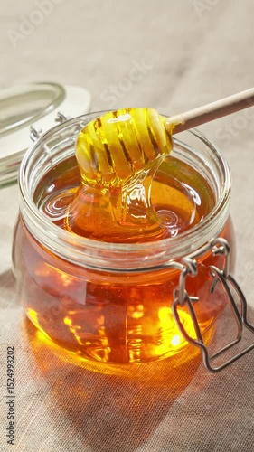 Jar filled with glistening honey sits atop soft linen surface. Wooden dipper swirls through golden liquid. Vertical video. Healthy organic honey. Honey with honey dipper in glass jar