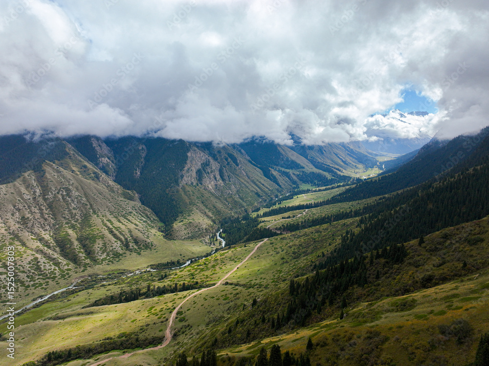 Fototapeta premium A sweeping aerial view captures a vast mountain valley with a river winding through its base, a dirt road on the slopes, and dramatic clouds overhead
