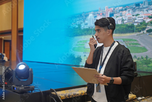 Asian male stage manager directs the show on stage using a headset, walkie-talkie, and clipboard, showing the professional work atmosphere behind the scenes of a show production.