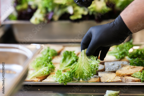 Chef with black gloves placing fresh lettuce on slices of bread in a professional kitchen, preparing sandwiches with hygiene and precision.