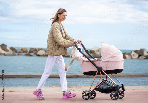 Full body young mother walks peacefully along a seaside promenade, pushing a pink baby stroller with a smile