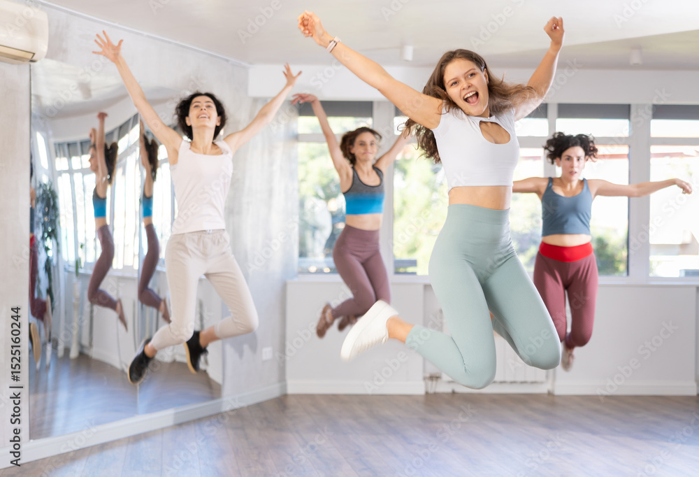 Fototapeta premium Cheerful sporty young girl captured in air while jumping during lively dance fitness routine with group of women in studio
