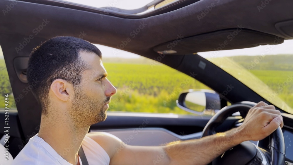 Profile of young handsome man driving a modern electric car with sunlight at background. Young male driver riding on electrical vehicle at countryside. Concept of journey and trip. Close up Profile of young handsome man driving a modern electric car with sunlight at background. Young male driver riding on electrical vehicle at countryside. Concept of journey and trip. Close up