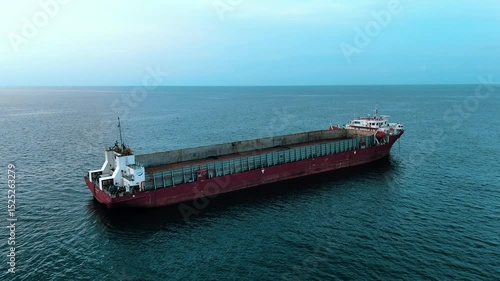 A large empty red barge or cargo ship sits alone on a calm open sea under a pale blue sky