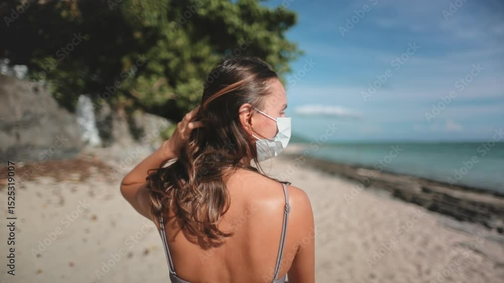Young woman is putting on a protective face mask while standing on a tropical beach. She is enjoying a sunny day by the sea but is aware of the importance of protecting herself and others Young woman is putting on a protective face mask while standing on a tropical beach. She is enjoying a sunny day by the sea but is aware of the importance of protecting herself and others