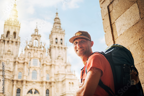 Joyful smiling middle-aged backpacker pilgrim sitting on Obradoiro square in Santiago de Compostela, Spain, with Santiago de Compostela Cathedral — famous Camino de Santiago finish point.