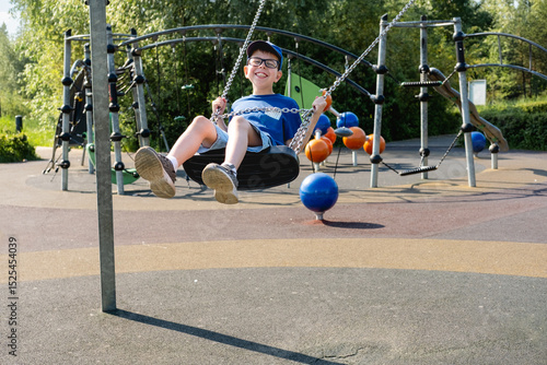 An 11 year old boy wearing glasses and a blue T-shirt is riding a swing in a park on a playground