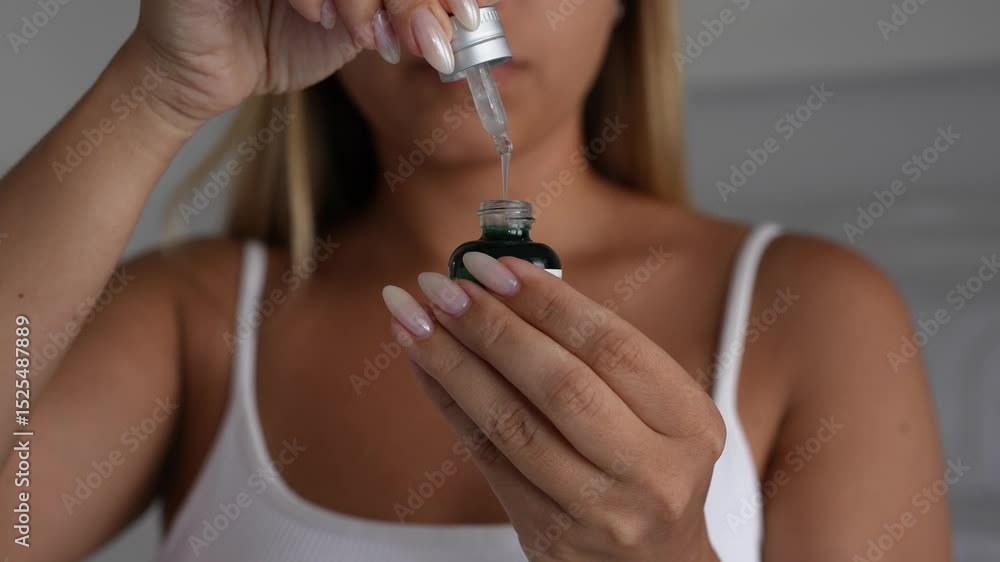 Young blonde woman squeezing moisturizing serum from a dropper into a glass bottle. Close-up beauty shot showing skincare ritual and glossy natural nails