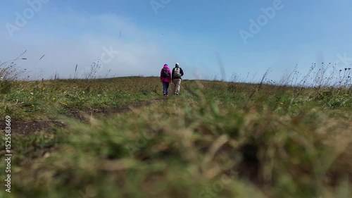  Two Backpackers - Haikers Walking Away on a Dirt Road, Ground-Level Shot, Rear View, Against a Blue Sky