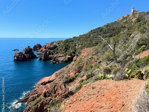 Coastal landscape in the Esterel region of southern France with dramatic red cliffs, deep blue sea, rugged rocks, and a distinctive observation tower perched on the hilltop.
