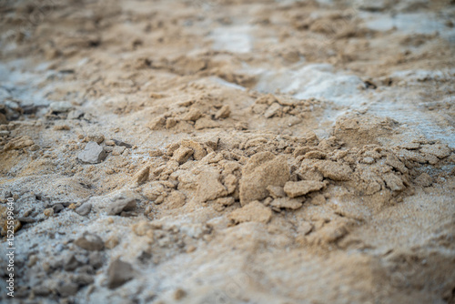 A close-up shot of textured sand, mixed with some small rocks and lighter material, fills the frame. The focus is on the details of the sands surface.