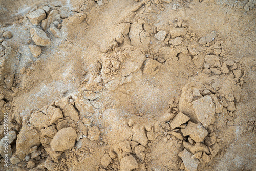 A close-up shot of textured sand, mixed with some small rocks and lighter material, fills the frame. The focus is on the details of the sands surface.