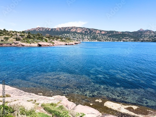 Clear coastal view of the Esterel mountains and turquoise Mediterranean Sea in southern France, with rocky shoreline and transparent water under a bright blue sky.

