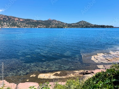 Clear coastal view of the French Riviera with deep blue sea, transparent water, coastal vegetation and distant hills under a bright blue sky.

