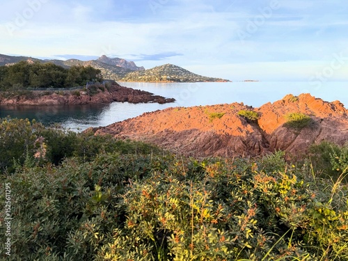 Golden hour light over the red rocky coastline of the Côte d’Azur, France, with the Esterel mountains in the background and calm Mediterranean waters.  
