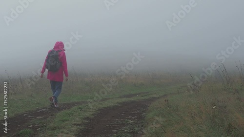 A woman in a red jacket with a backpack is walking away from the camera on a dirt road in the mountains. The scene is set in strong wind, surrounded by clouds and mist. The atmosphere is windy, rugged