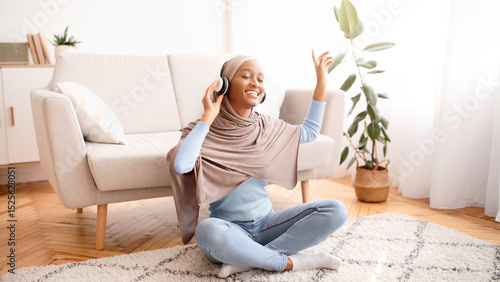 Obraz na plátně Positive black woman in hijab sitting on floor, wearing headphones, listening to music at home