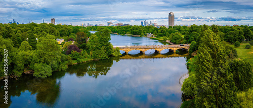 Aerial view of Hyde Park in London featuring the Serpentine lake, lush greenery, and a stone bridge with city skyline in the background