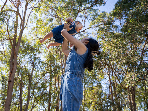 Mother lifting baby joyfully in a forest under clear blue sky