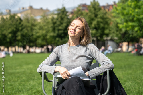 Bilde på lerret Content woman squinting while enjoying summer sunshine, relaxing in park after work seated on chair with documents
