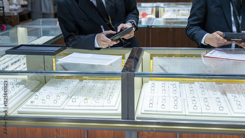 Photos Two jewelry store clerks in formal uniforms inspecting ring displays, using a tablet and clipboard to record inventory inside a brightly lit showcase counter