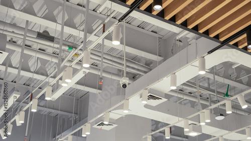 Photos Modern commercial ceiling with industrial-style exposed pipes, hanging LED lights, CCTV camera, air ducts, and fire sprinkler system