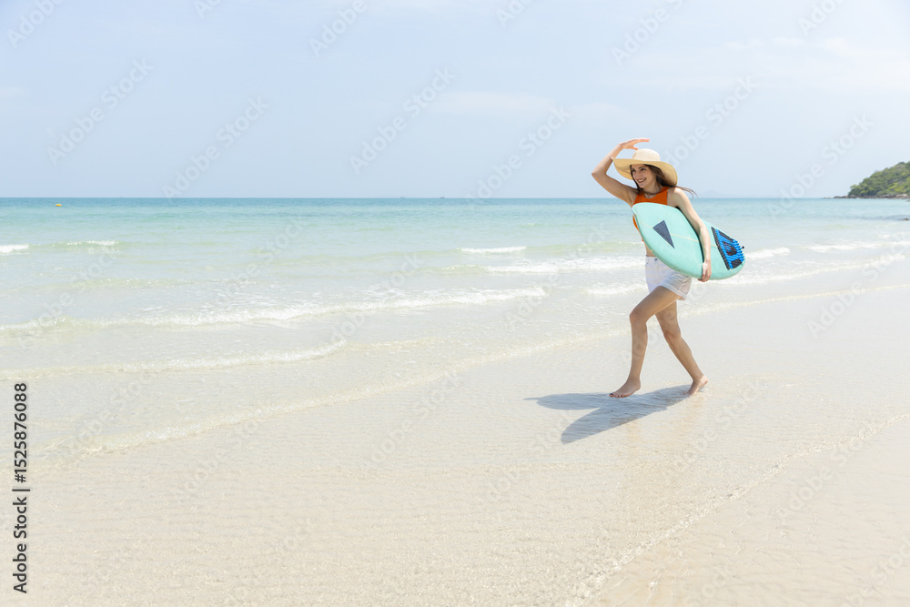 Joyful young woman walks out of water at sunny beach, holding surfboard. She wears bright orange top and white shorts, with clear blue sky in background. Joyful young woman walks out of water at sunny beach, holding surfboard. She wears bright orange top and white shorts, with clear blue sky in background.
