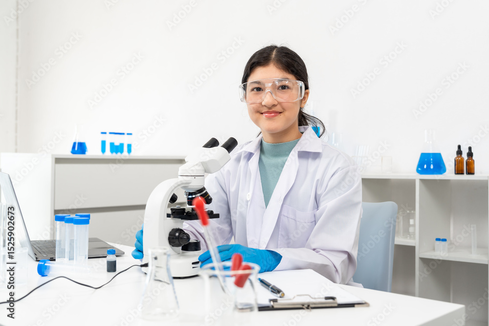 Asian female teenager in lab coat and goggles smiling confidently while seated beside microscope in science classroom. Surrounded by test tubes, beakers, clipboard and laptop on clean white desk Asian female teenager in lab coat and goggles smiling confidently while seated beside microscope in science classroom. Surrounded by test tubes, beakers, clipboard and laptop on clean white desk