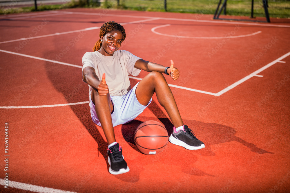 Obraz premium Young black woman practicing basketball alone at sports court in the morning. Happy girl sitting on basketball court on sunny day.