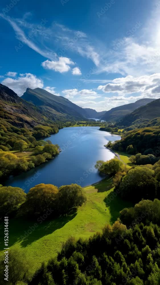 Scenic valley landscape with blue lake reflecting sky, green grassy hills, and mountain peaks under cumulus clouds Scenic valley landscape with blue lake reflecting sky, green grassy hills, and mountain peaks under cumulus clouds