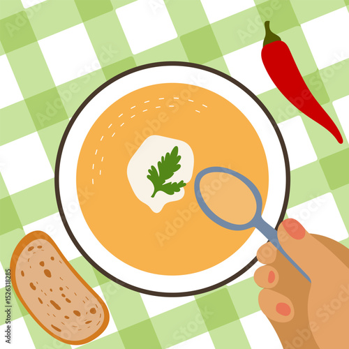 Pumpkin soup with sour cream and parsley. Someone is eating a tasty dish with a spoon. A plate with a delicacy on a tablecloth. A piece of bread and chili pepper