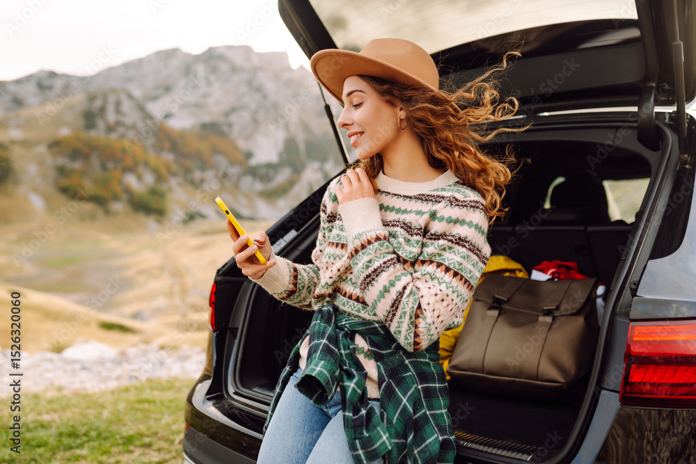 Naklejka premium Beautiful woman in the trunk of a car with a phone enjoying a road trip. Young traveler blogging and observing the mountain scenery from the car. Wanderlust.