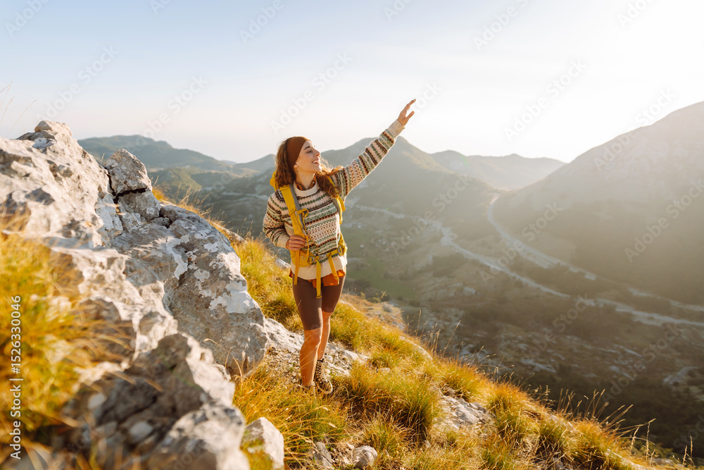 Obraz premium A young female traveler with a colorful hiking backpack walks along a mountain peak. A cheerful woman feels free and enjoys the mountain landscape at sunset. Hiking concept. Active lifestyle.