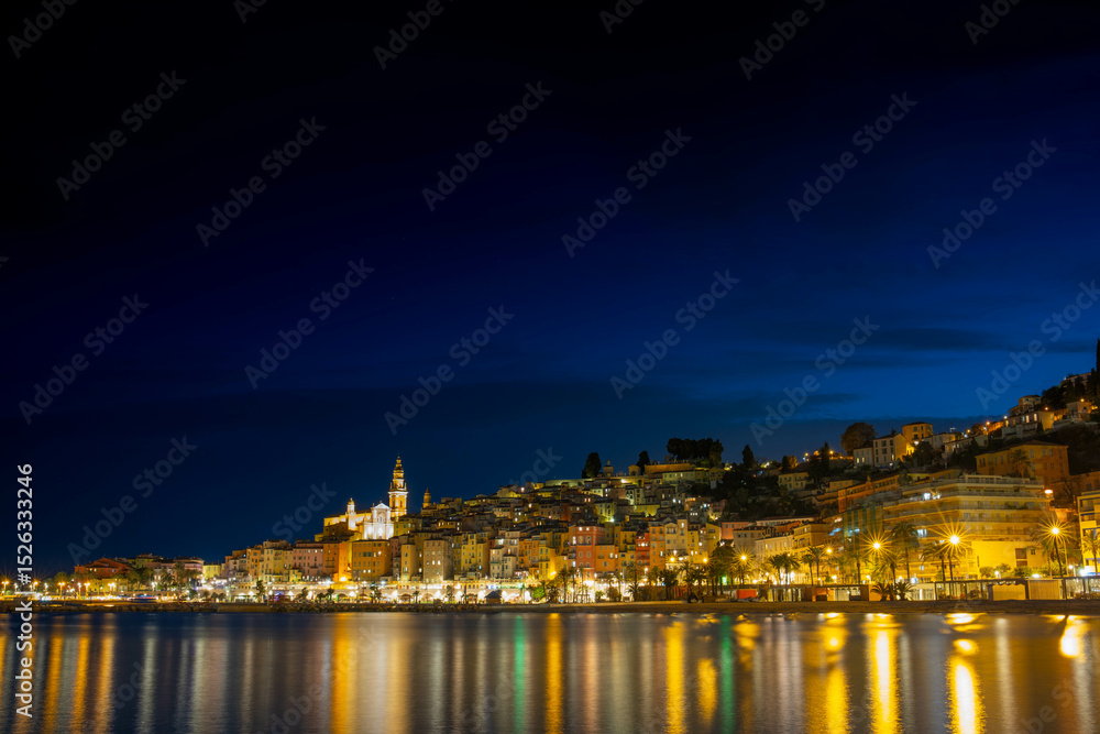 Fototapeta premium Night panorama of Menton in France. The lights of the night city are reflected on the calm sea surface.