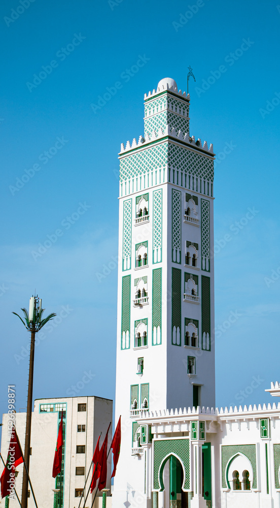 Obraz premium Elegant white and green minaret of a mosque in Tetouan, Morocco, under a clear blue sky, surrounded by red Moroccan flags.