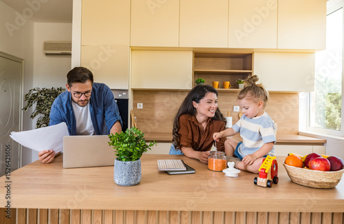 Fototapet Joyful young family bonding in bright modern kitchen mother and daughter laughing while playing on countertop as father focuses on remote work, blending parenthood, love, and technology