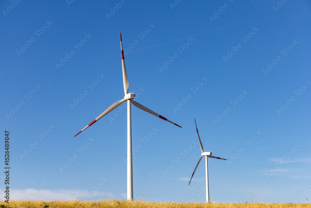 Fototapeta premium Wind turbines in an open countryside field under a clear blue sky, used for renewable energy production and clean power generation.